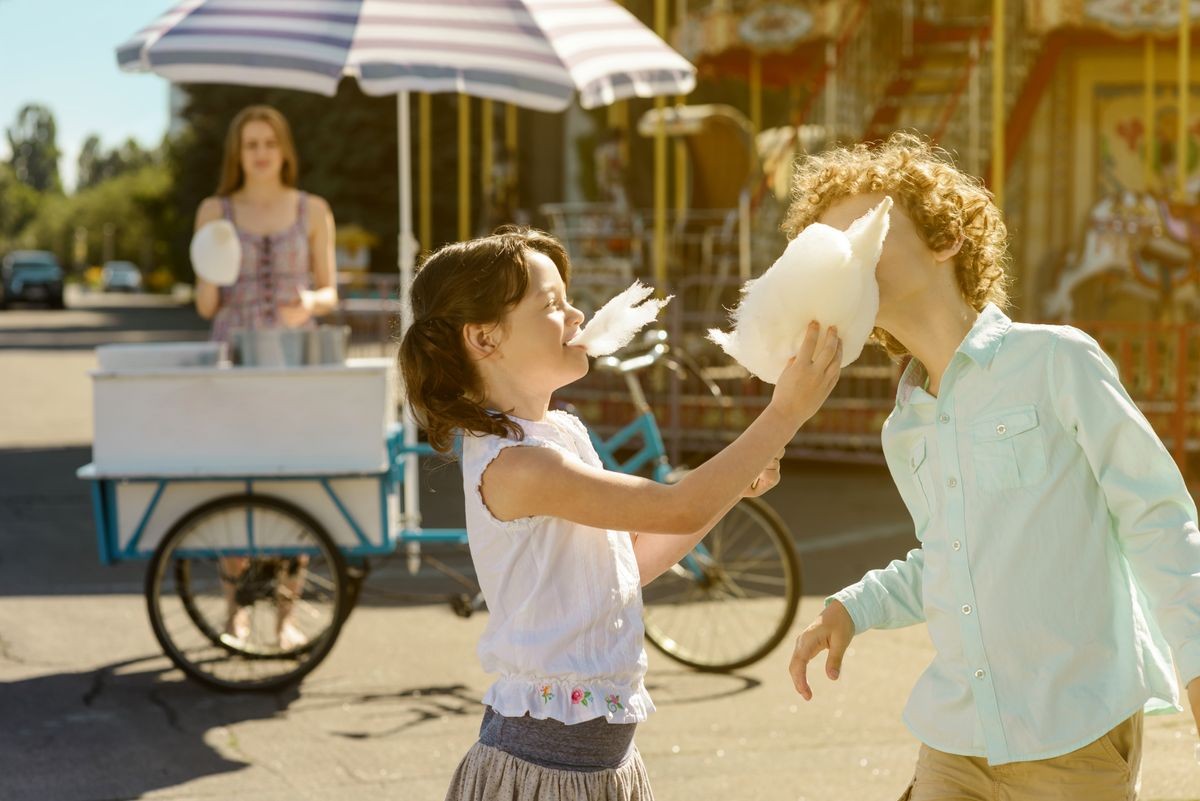 Funny kids eat cotton candy. One dessert for two little close friends. Sales point with blue bicycle and attractive worker in the background.